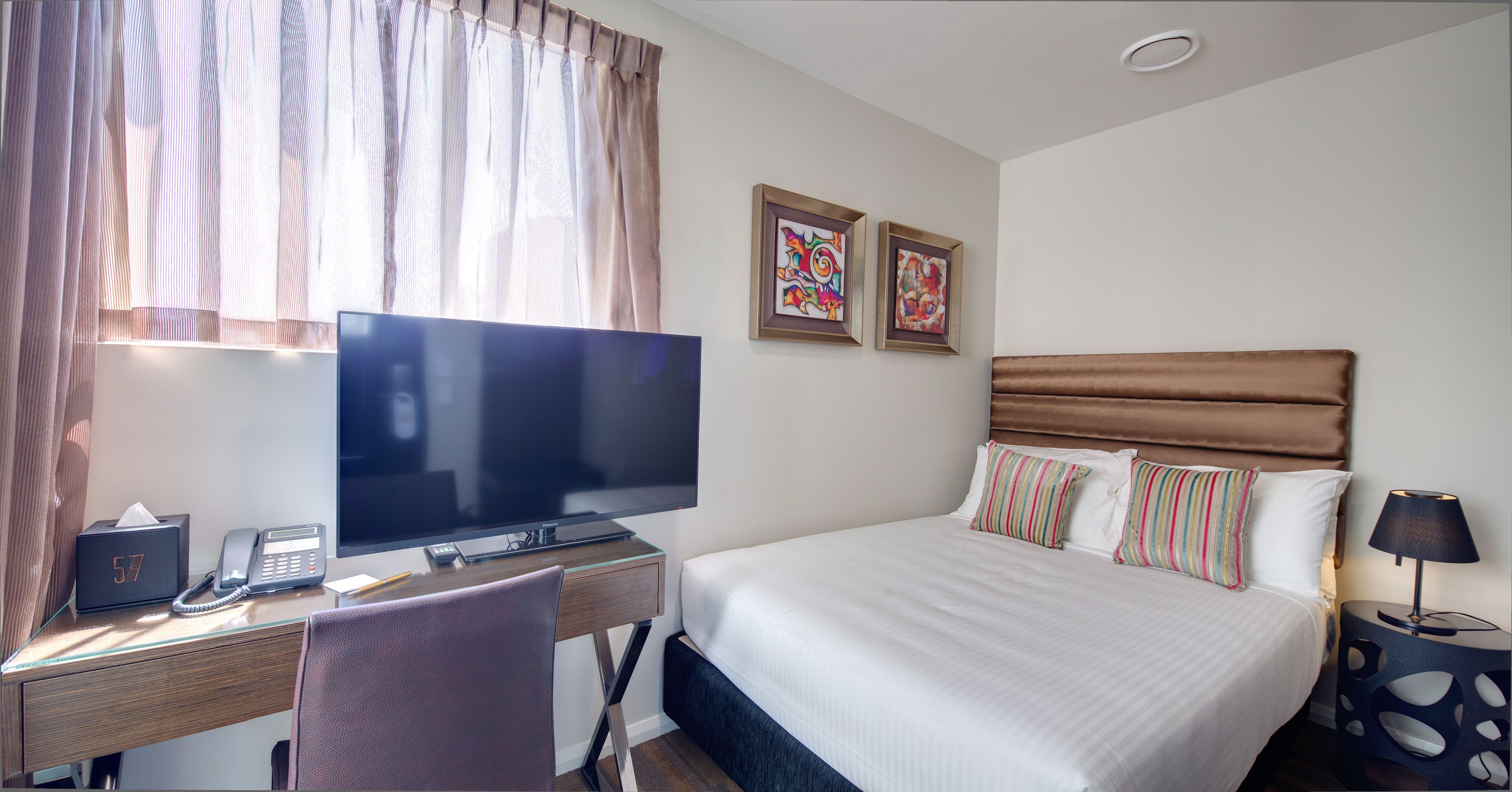 A hotel room with a bed, a desk with a computer, a telephone, and two framed artworks on the wall.