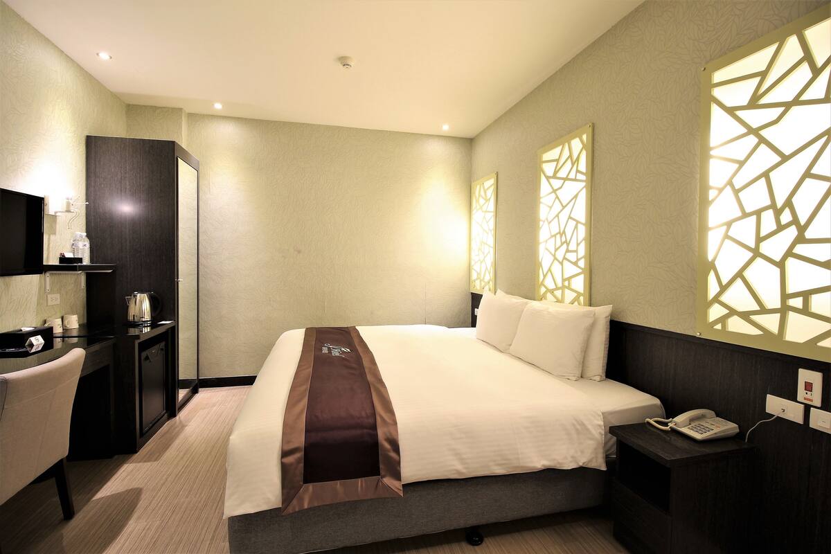 A hotel room with a bed, bedside table with a phone, a desk with a chair, and a TV.