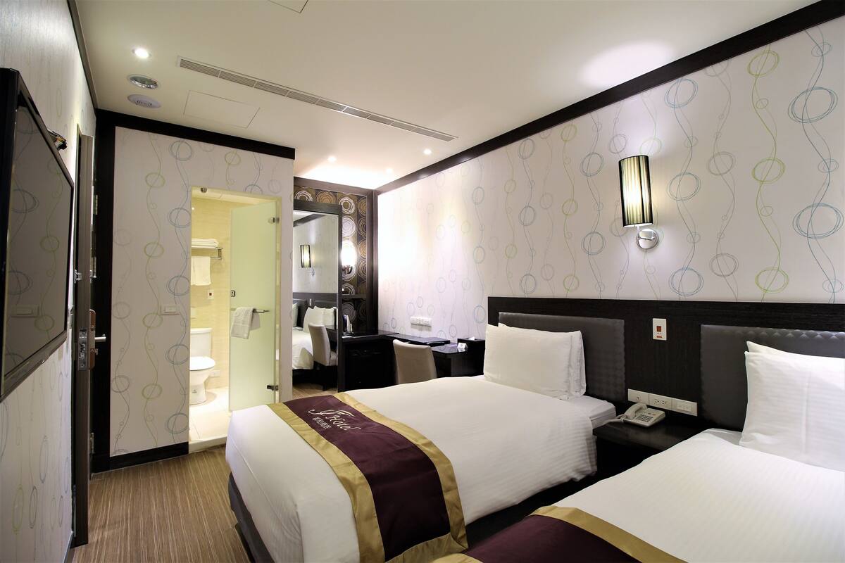 A hotel room with two beds, a desk, a chair, a bathroom, and a mirror.