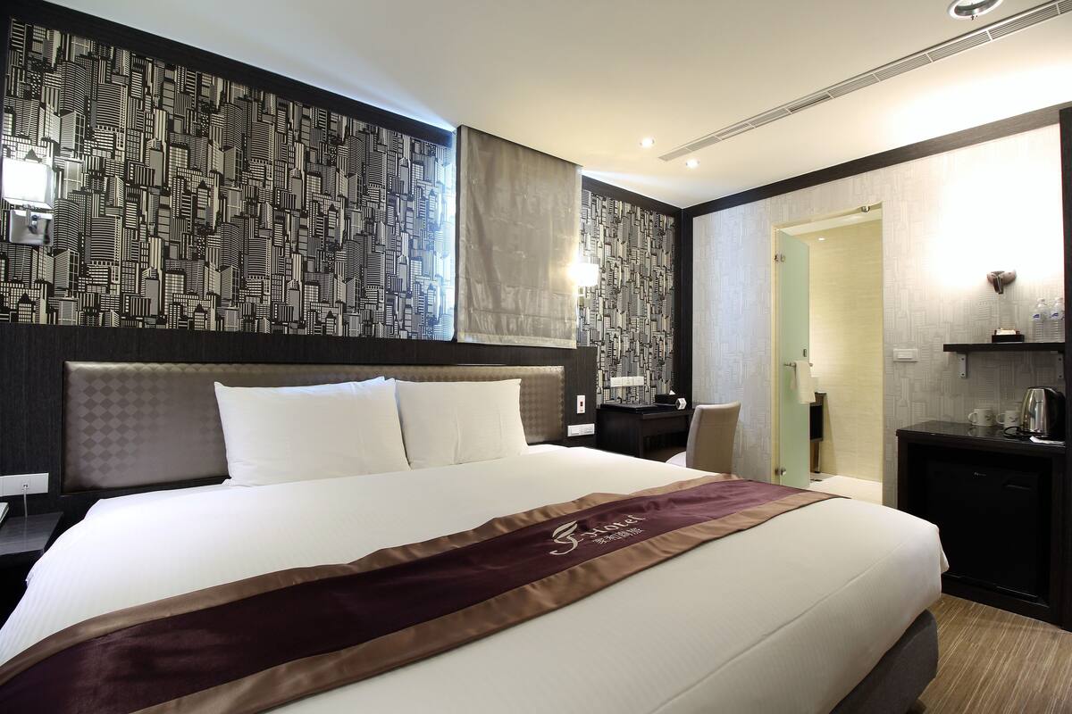 A hotel room with a large bed, a headboard with a geometric pattern, a bedside table with a lamp, and a mirror.