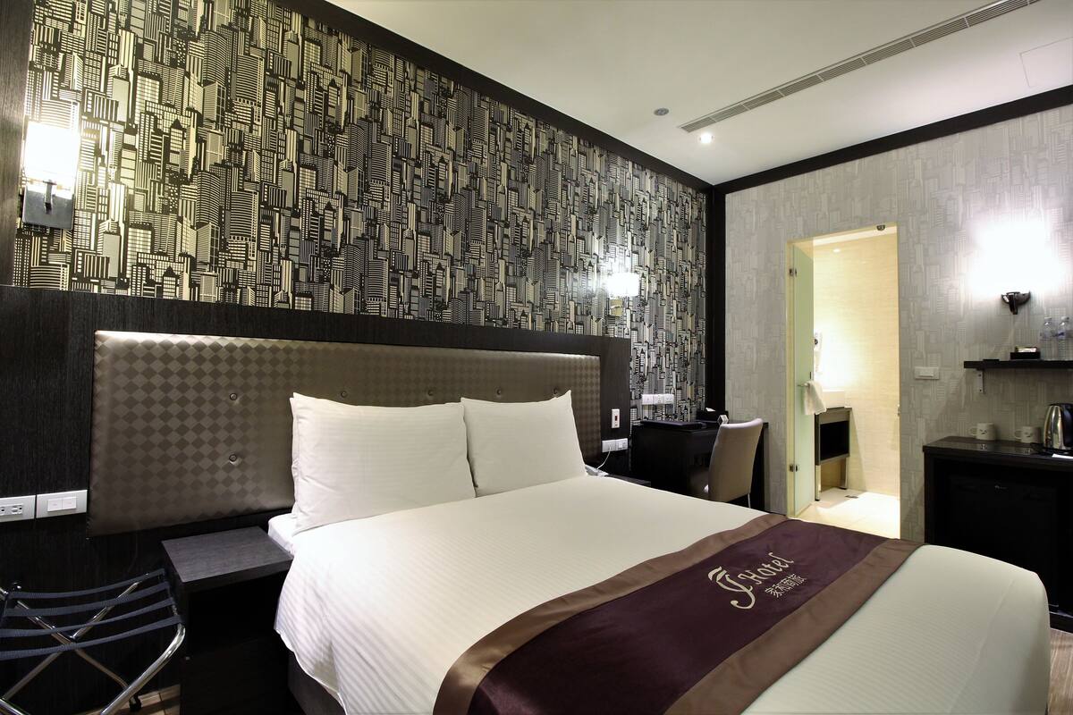 A hotel room with a large bed, a headboard with a geometric pattern, a desk with a chair, and a small kitchenette area.