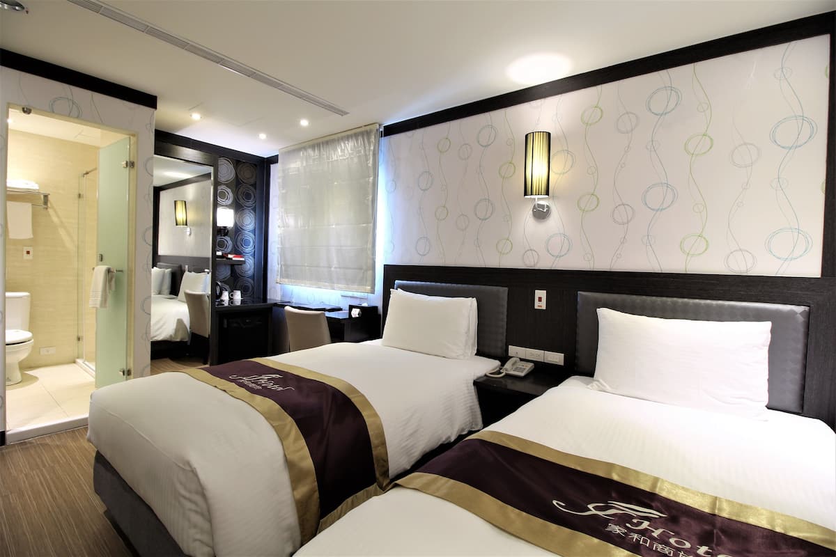 A hotel room with two beds, a desk, a mirror, and a shower area.