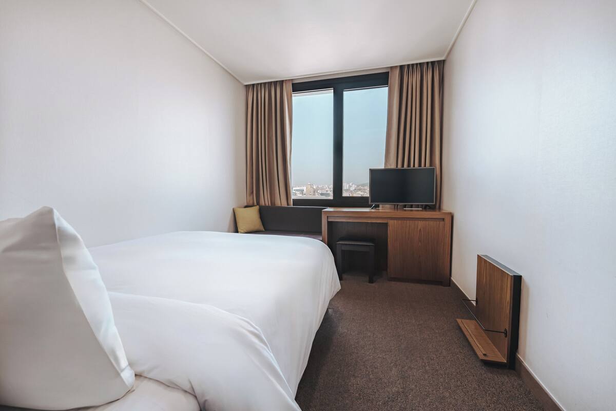 A hotel room with a bed, a desk, a television, and a view of the city through large windows.