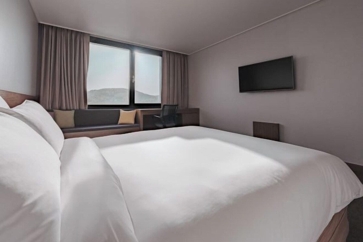 Habitació d'hotel amb un llit gran, una finestra amb cortines, un televisor de pantalla plana fixat a la paret i una zona d'estar amb una cadira i una taula.