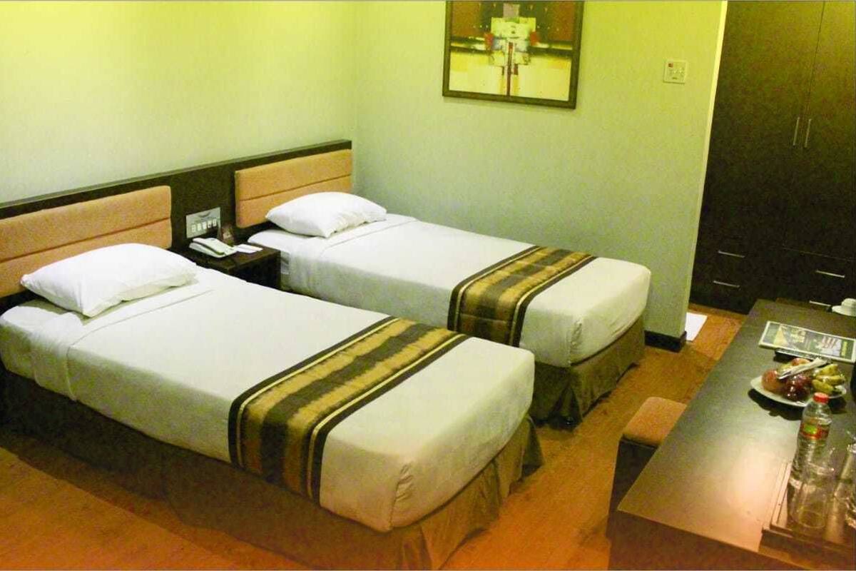 Habitación de hotel con dos camas individuales, una mesita de noche con un teléfono, una pequeña mesa con una fuente de fruta y una botella, y un espejo en la pared.