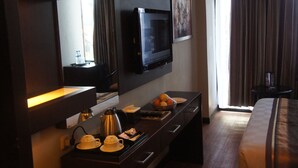 Quarto de hotel com TV de tela plana na parede, mesa de cabeceira com chaleira, xícaras e uma fruteira, e uma cama com colcha xadrez.