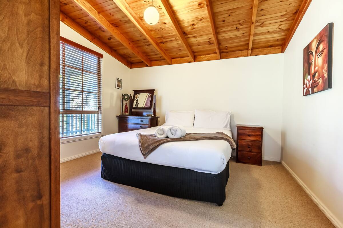 Ein Schlafzimmer mit Holzboden, einem großen Bett, einem Nachttisch, einer Kommode und einem Fenster mit Jalousien.