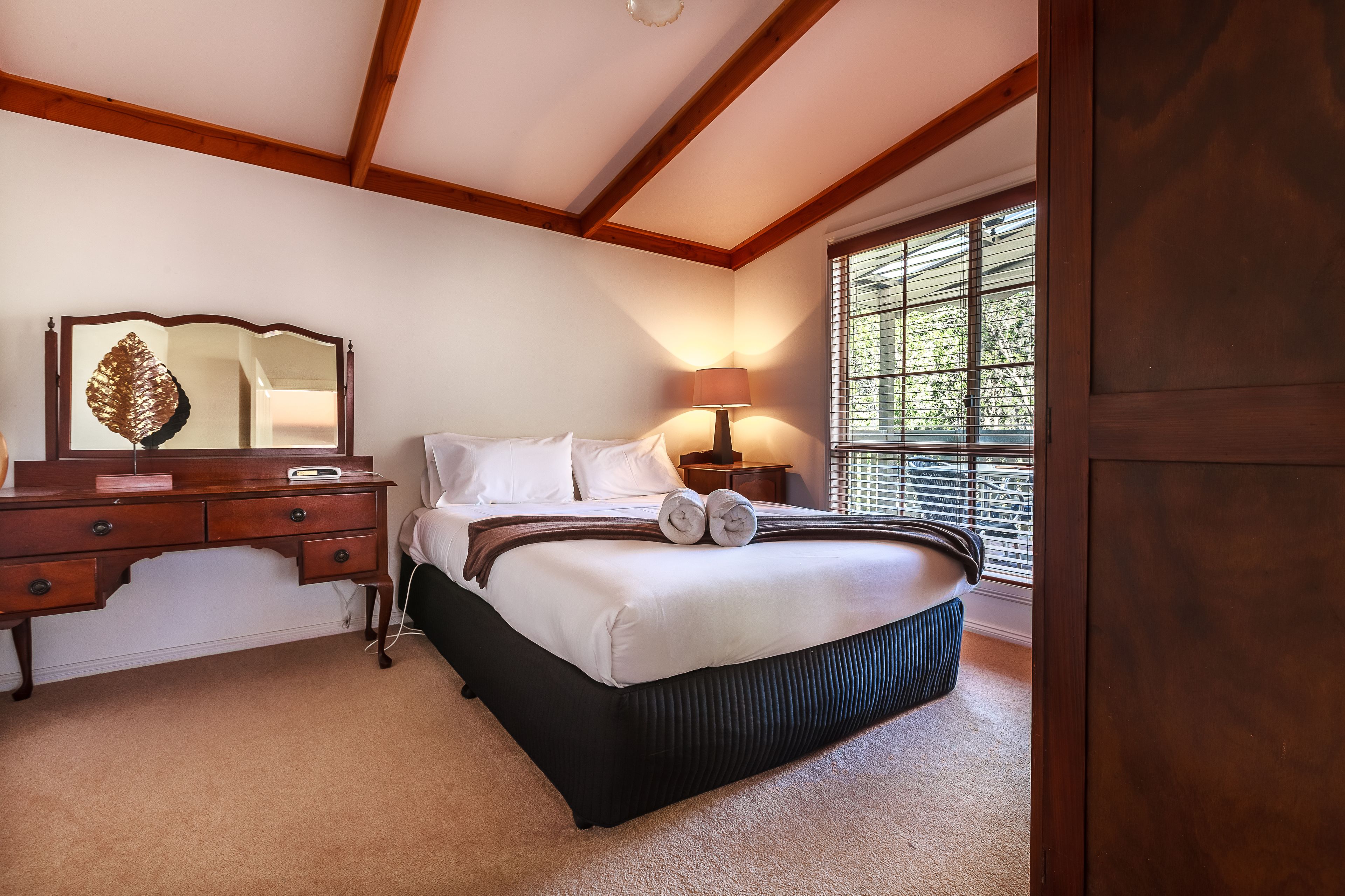 A bedroom with a large bed, a wooden dresser, a mirror, and a window with blinds.
