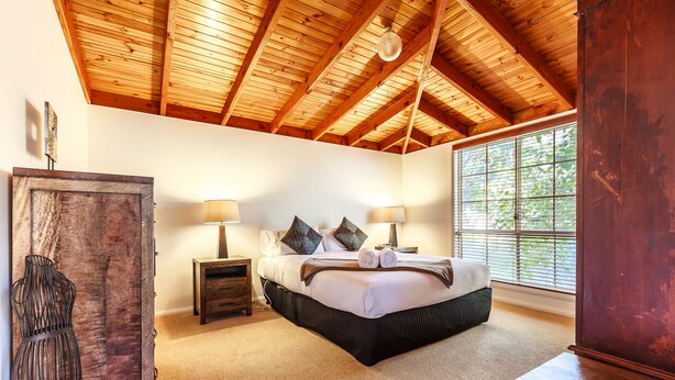 Un dormitorio con techo de madera, un ventanal grande, una cama con almohadas, una mesita de noche y un aparador.