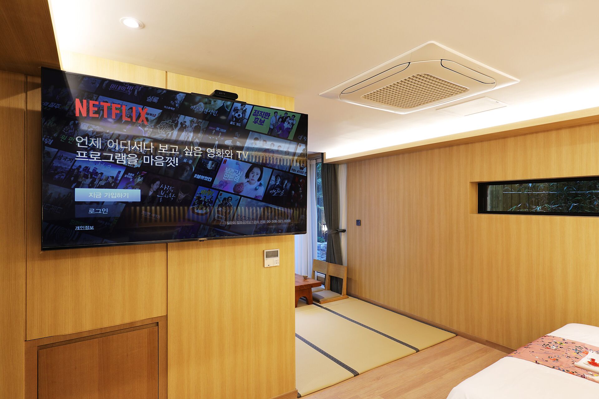 Habitación de hotel con una televisión grande de pared que muestra varias pantallas, detalles de madera, una pequeña mesa con una silla y una cama tipo futón.