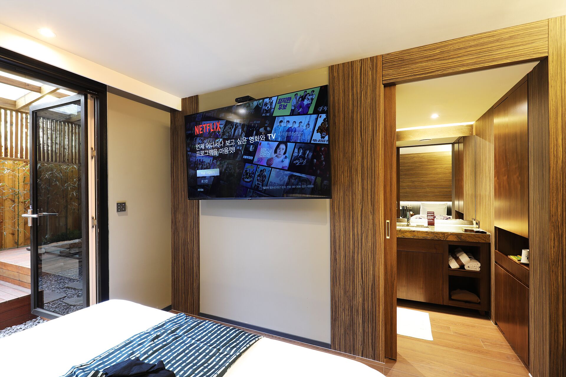 A modern hotel room with a large TV mounted on the wall displaying various content, a bed with a striped bedspread, wooden paneling, and a visible bathroom with a vanity and towel rack.