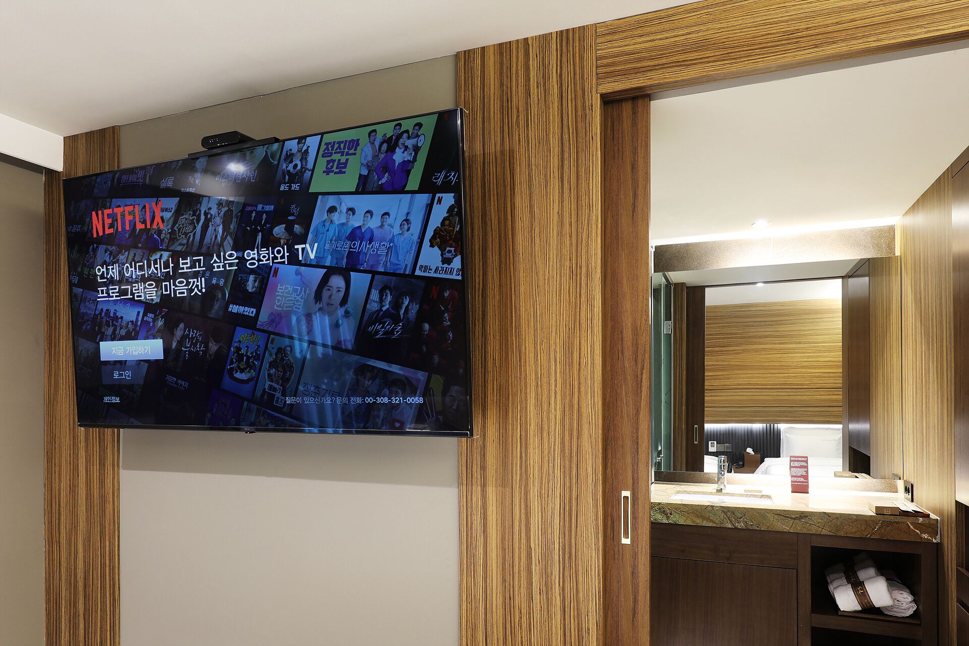 A hotel room with a wall-mounted TV displaying various Korean TV shows and a wooden panel design.