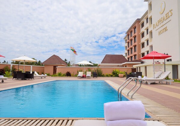 A swimming pool with lounge chairs and umbrellas, surrounded by a hotel building with flags. - Raphael's Hotel (Pemba)