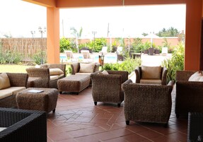 Un área de estar techada al aire libre con mobiliario de mimbre y una piscina al fondo.