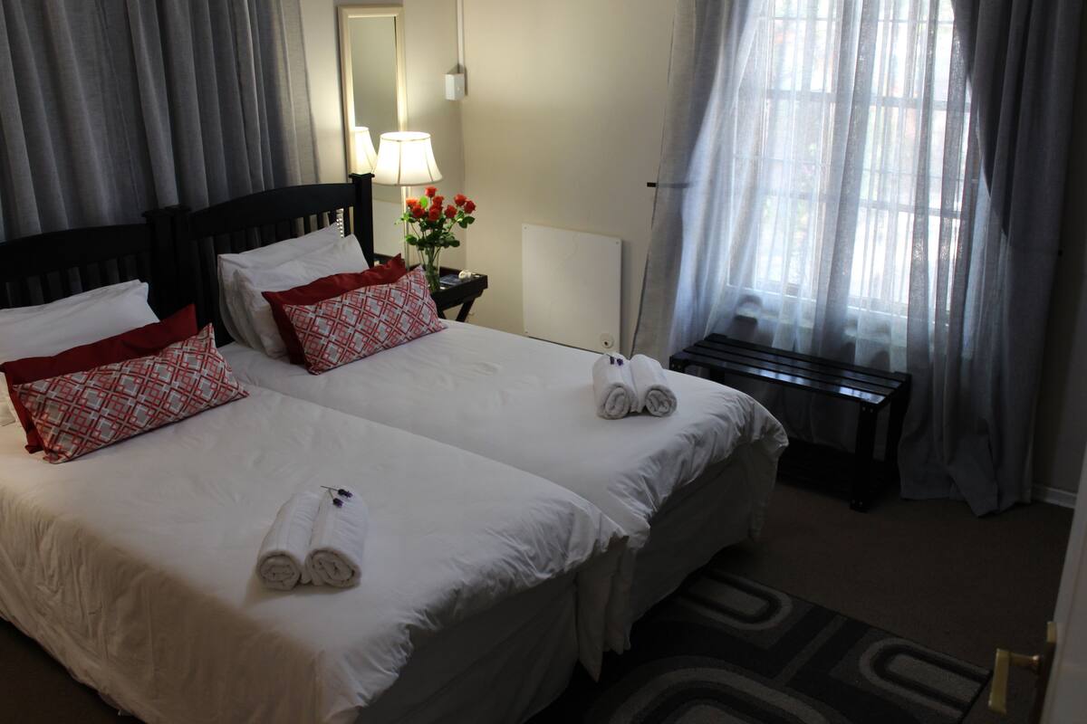 Ein Hotelzimmer mit zwei Betten, jeweils mit weißen Leinentüchern und roten Kissen. Es gibt einen kleinen Tisch mit einer Lampe und einer Blumenvase.