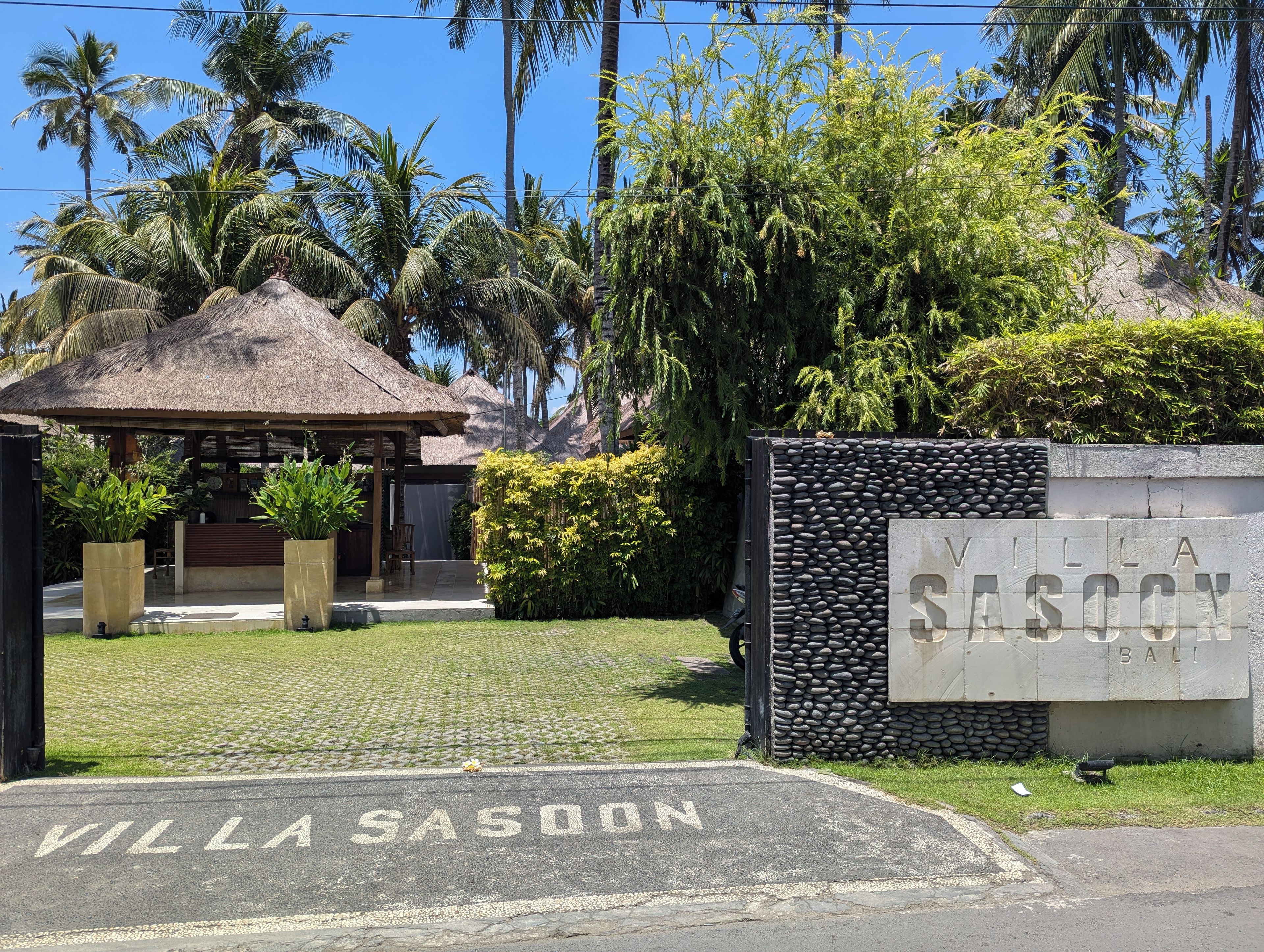 A villa entrance with a thatched roof, a stone wall, and a sign reading 'Villa Sasoon Bali'.