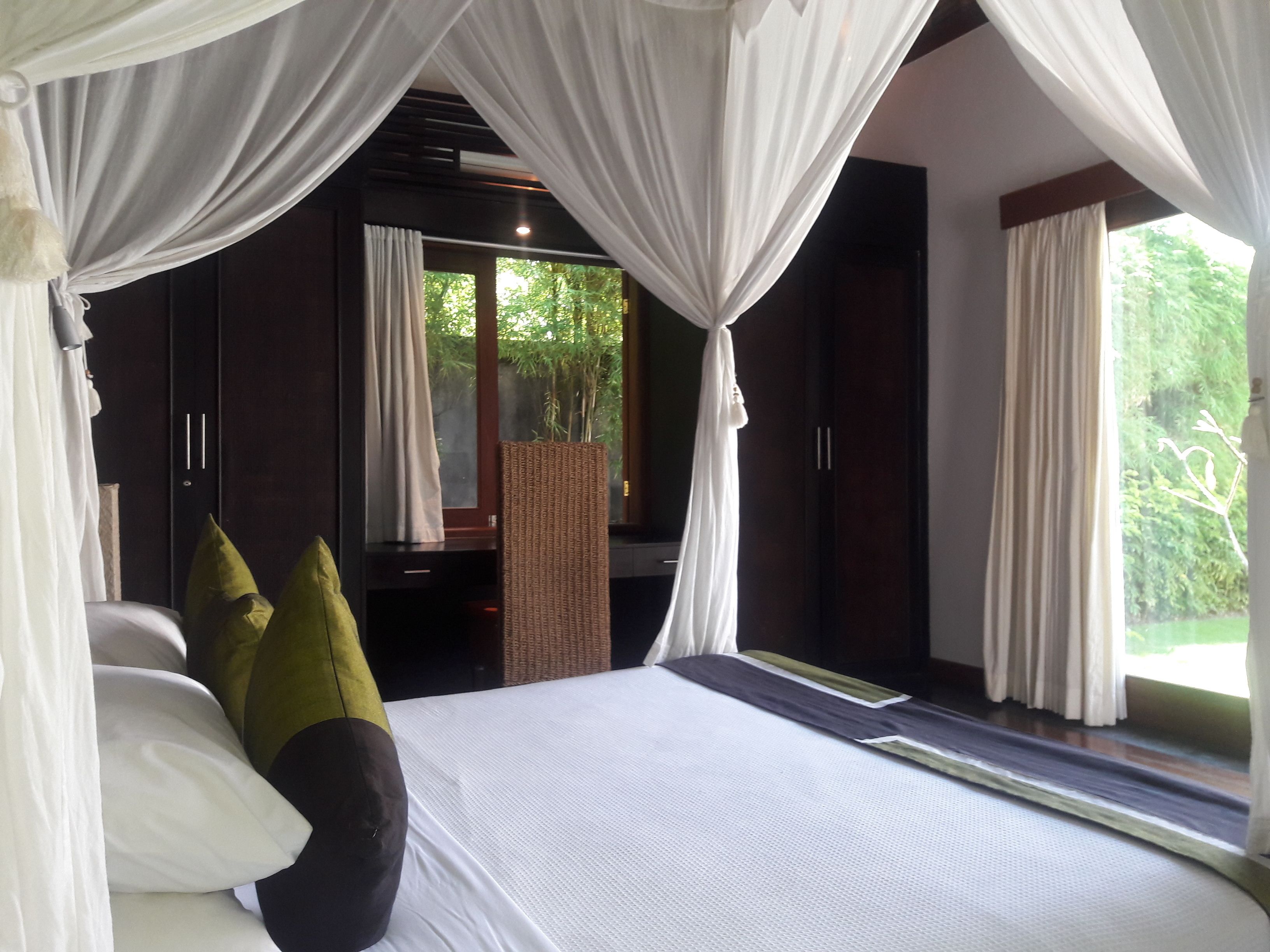 Quarto com cama, cortinas brancas e vista para o verde lá fora.