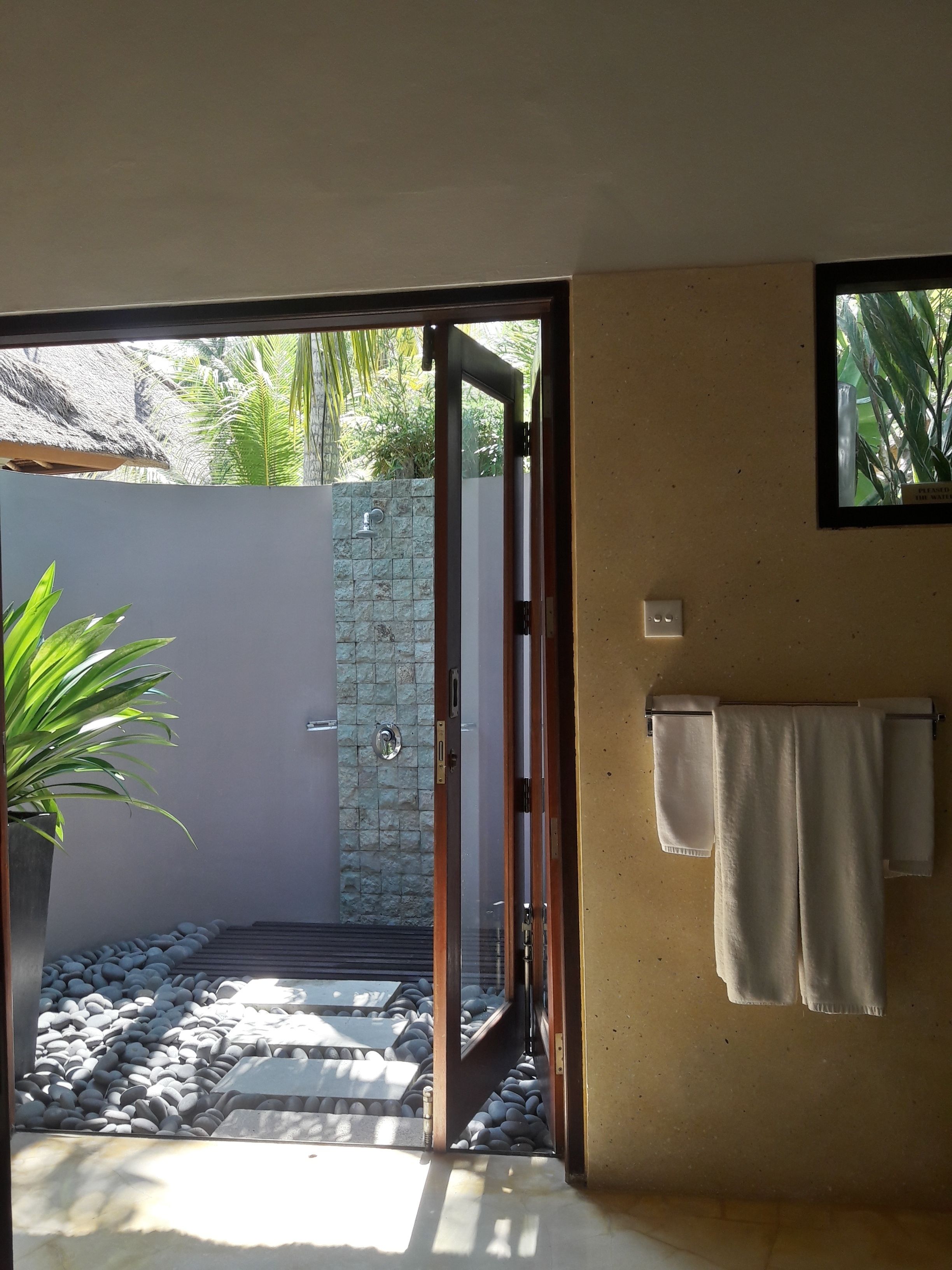 Un baño con puerta de cristal que da a un jard ín con muro de piedra, techo de paja y toallero con toallas.