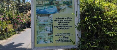 A signboard for Villa Sasoon, detailing amenities like two bedrooms, a large landscape, tropical garden, private pool, fully equipped kitchen, and free Wi-Fi.