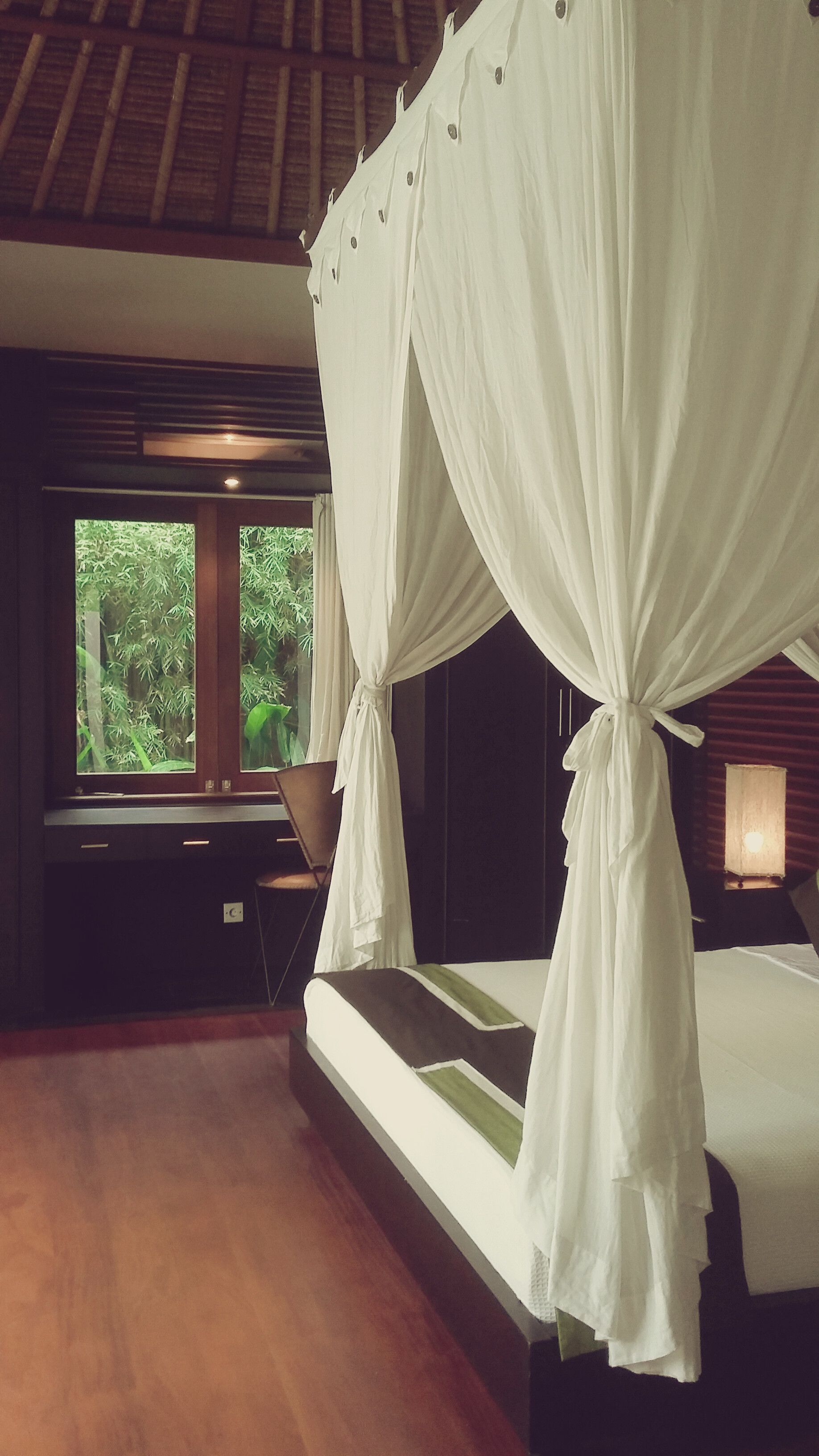 A bedroom with a four-poster bed, a large window with a view of greenery, and a wooden floor.
