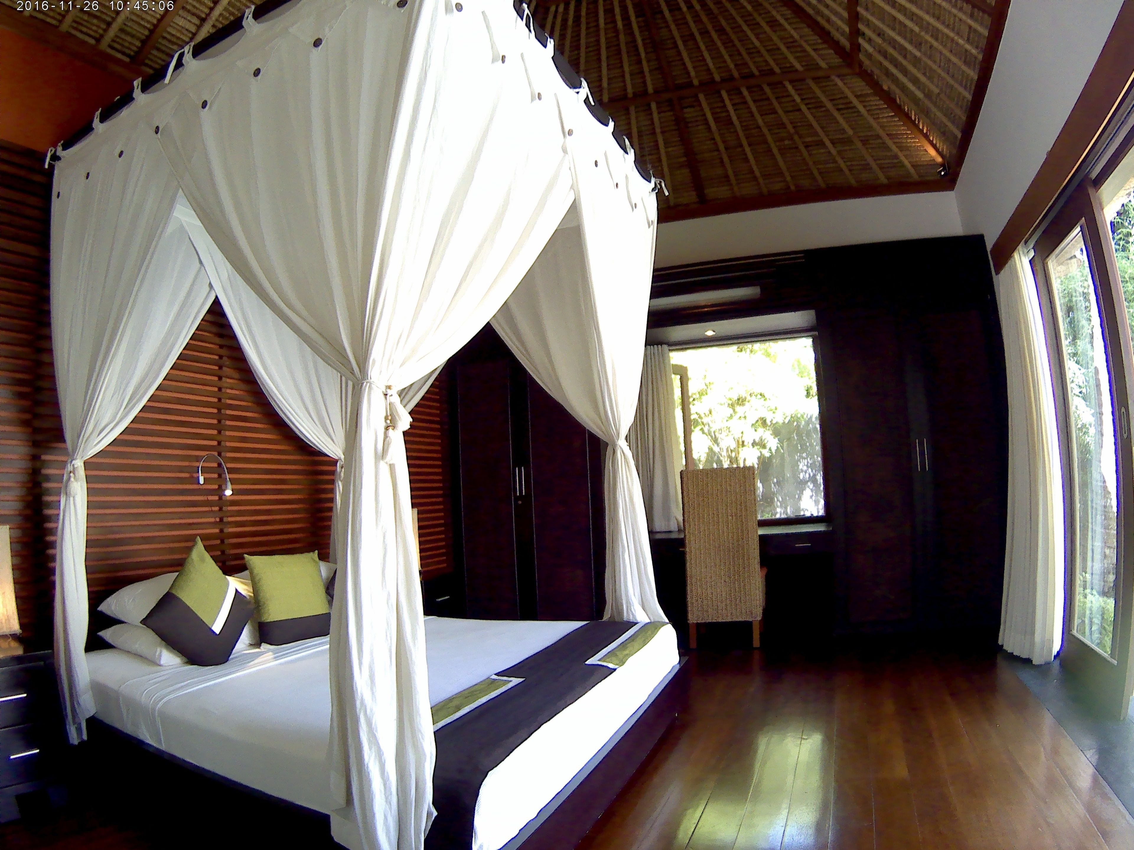 A four-poster bed with white drapes, wooden flooring, and a view of greenery outside.