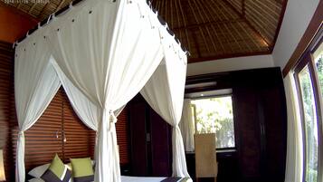 A four-poster bed with white drapes, wooden flooring, and a view of greenery outside.