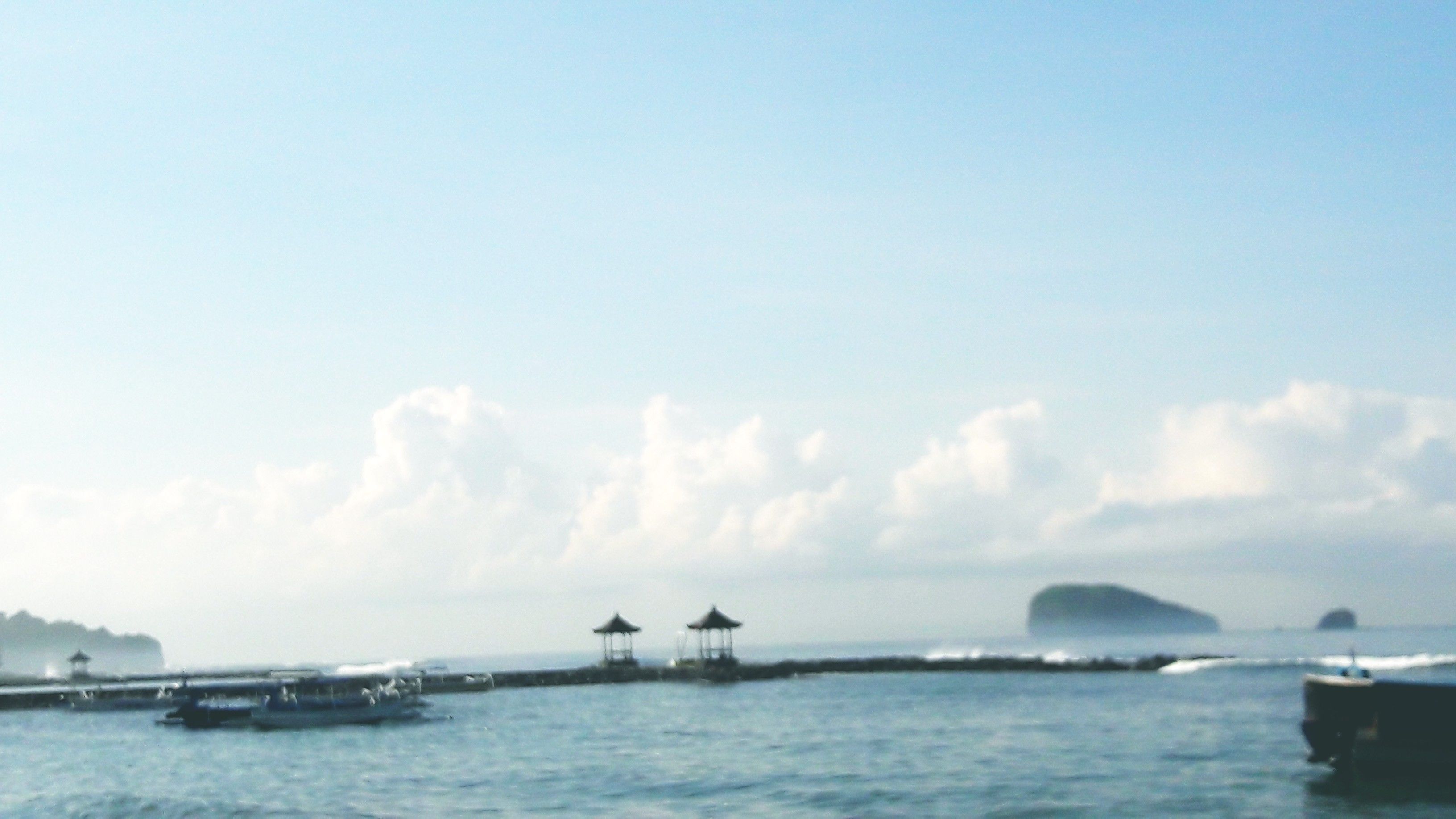 A coastal scene with a pier extending into the water, featuring several small structures. Boats are visible on the water, and a large island is seen in the distance.
