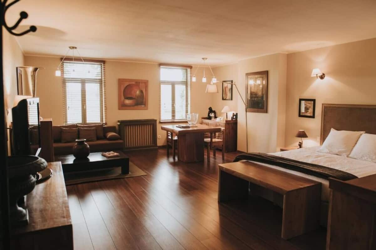 Een hotelkamer met een bed, een slaapbank, een eettafel en een salontafel. Er zijn twee ramen met jaloezieën, een radiator en een lamp aan de muur.