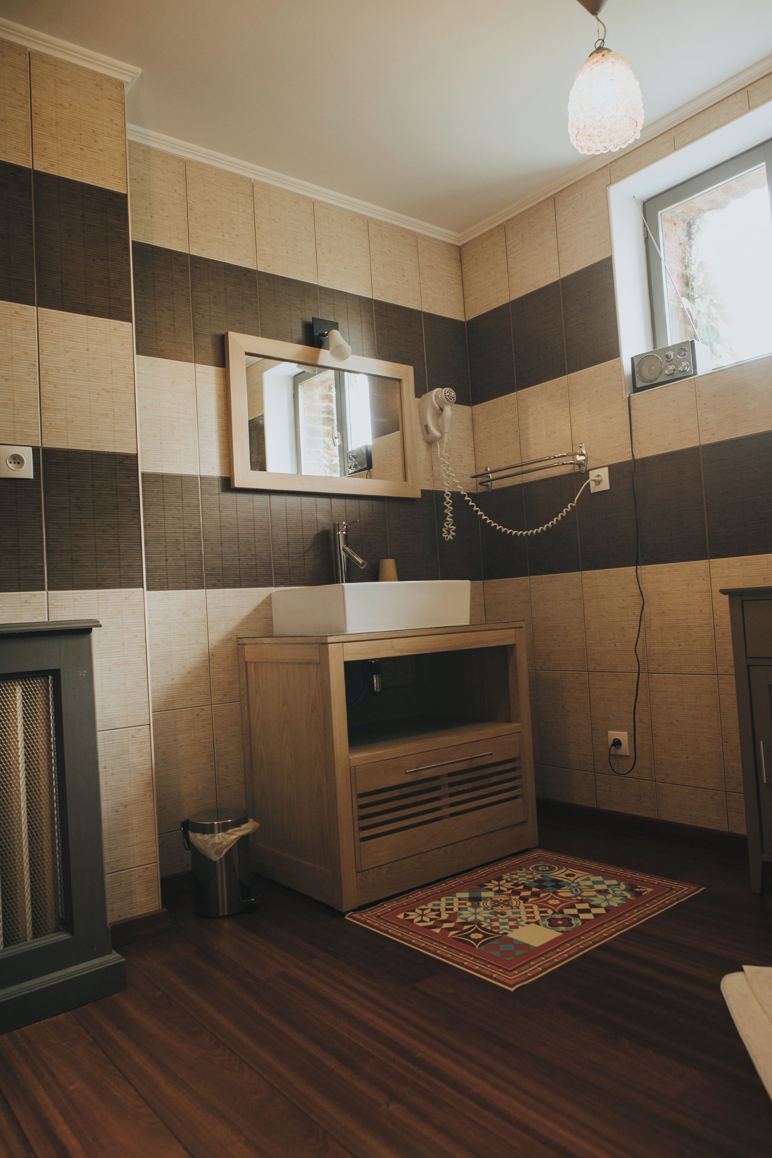 Un baño con tocador de madera, un lavamanos, un espejo, una lámpara colgante y un tapete estampado en el suelo.
