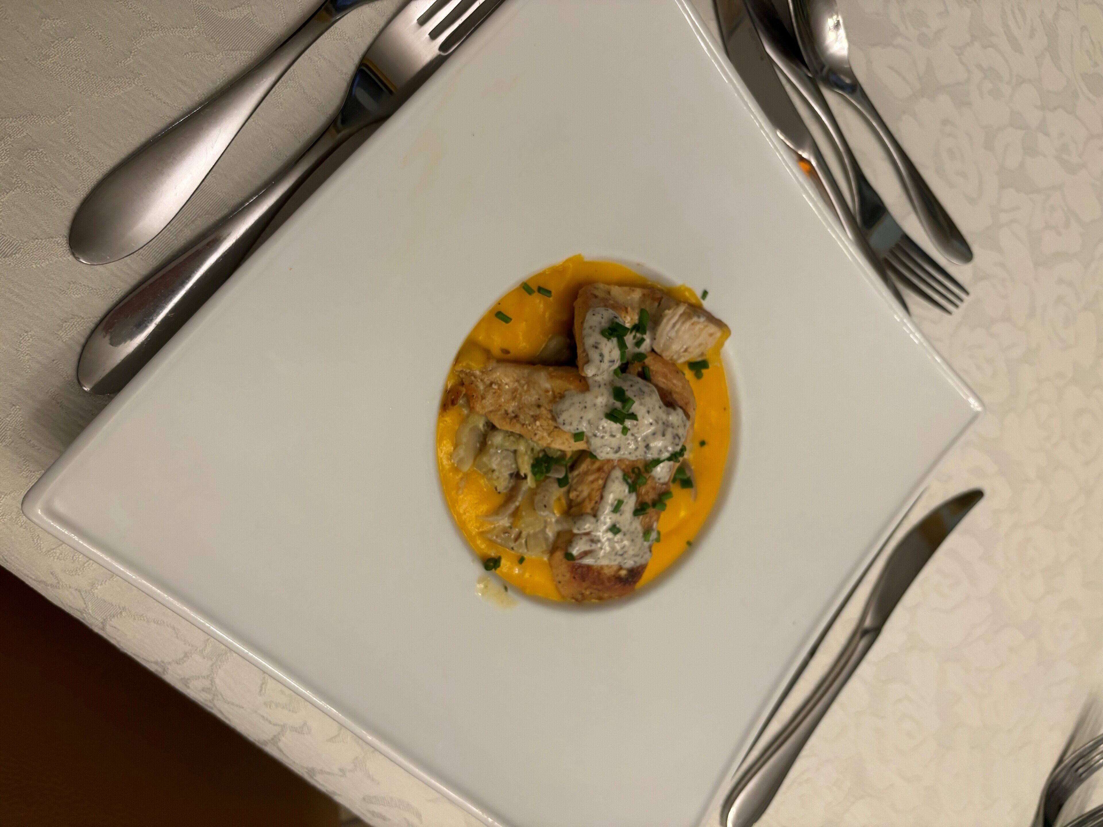 Ein weißes, quadratisches Teller mit Fleisch und Soße, dazu Besteck auf einer Tischdecke.