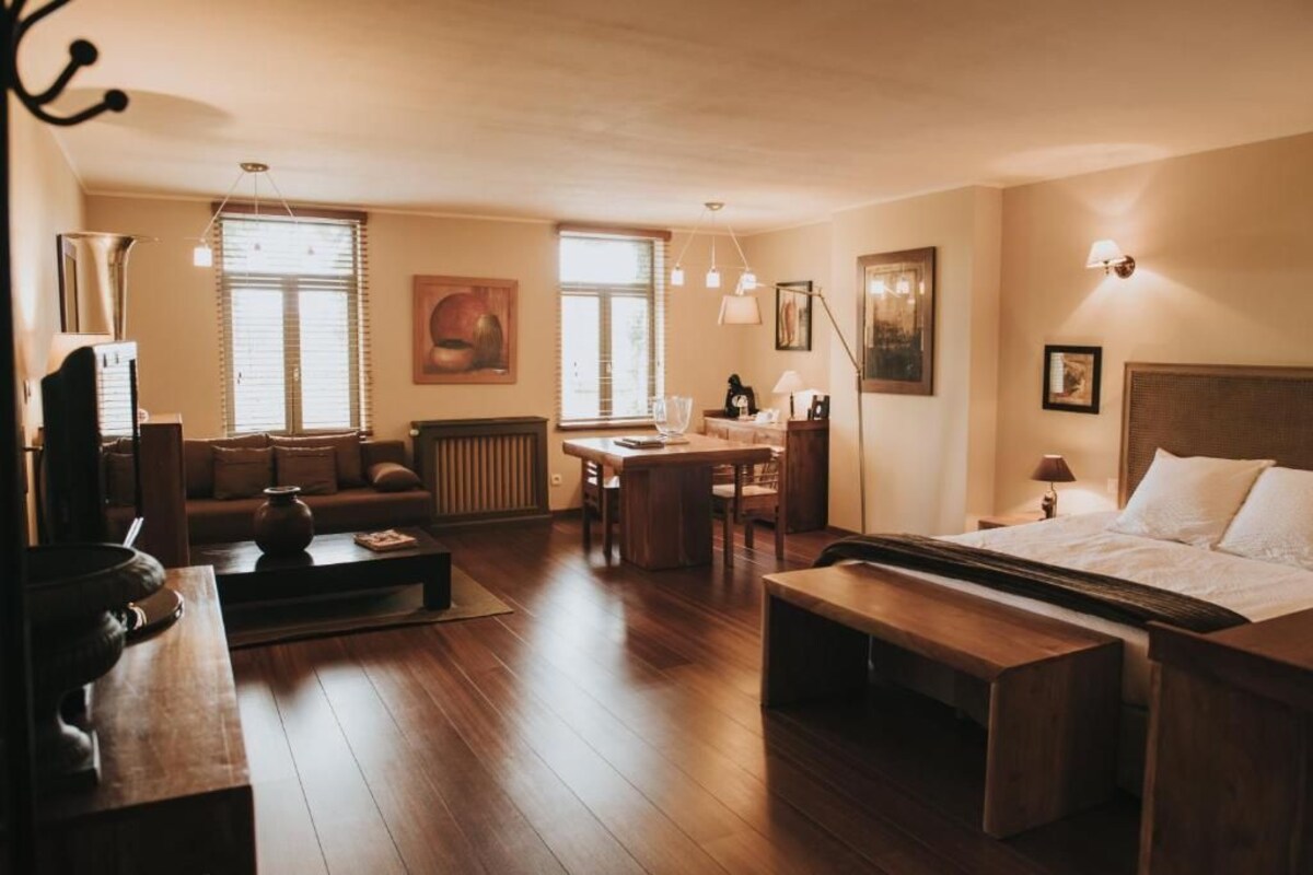 Ein Hotelzimmer mit Bett, Sofa, Esstisch und Couchtisch. Zwei Fenster mit Jalousien, ein Heizkörper und eine Wandlampe sind vorhanden.