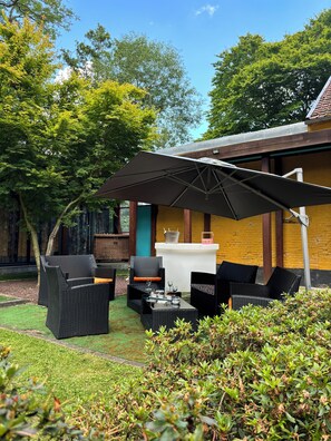 Een terras met rieten meubilair en een grote parasol, voor een geel gebouw met een bakstenen muur.