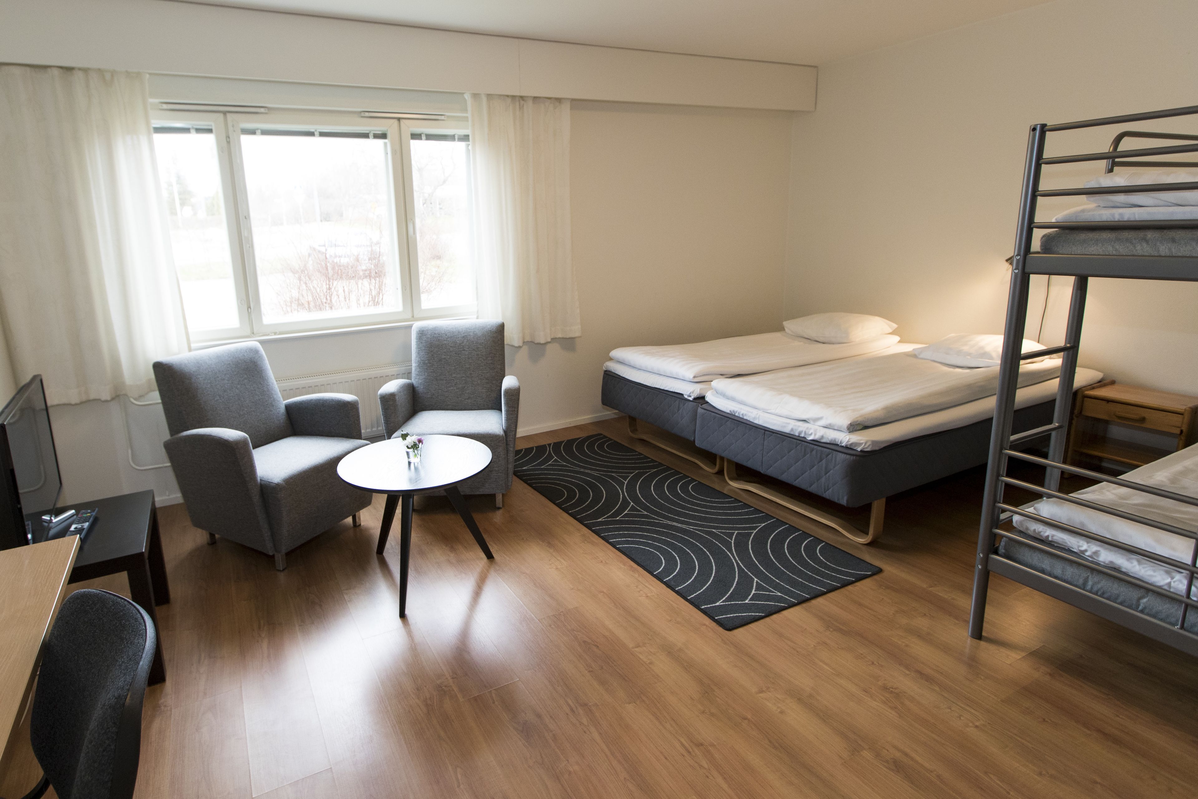 Ein kompaktes Hotelzimmer mit Etagenbett, kleinem Schreibtisch, Stuhl, rundem Tisch und einem Fenster mit Vorhängen.