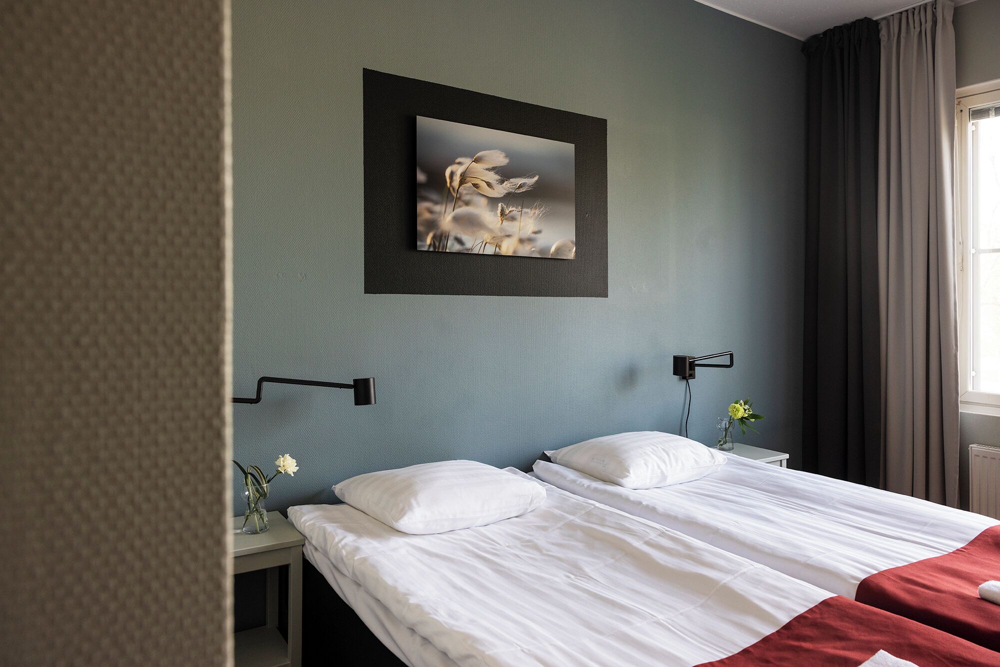 Una habitación de hotel con cama, mesita de noche, lámpara de pared, cuadro enmarcado y ventana con cortinas.