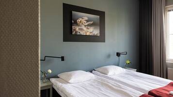 A hotel room with a bed, a nightstand, a wall-mounted lamp, a framed picture, and a window with curtains.