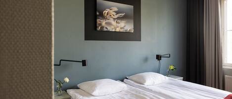 Una habitación de hotel con cama, mesita de noche, lámpara de pared, cuadro enmarcado y ventana con cortinas.