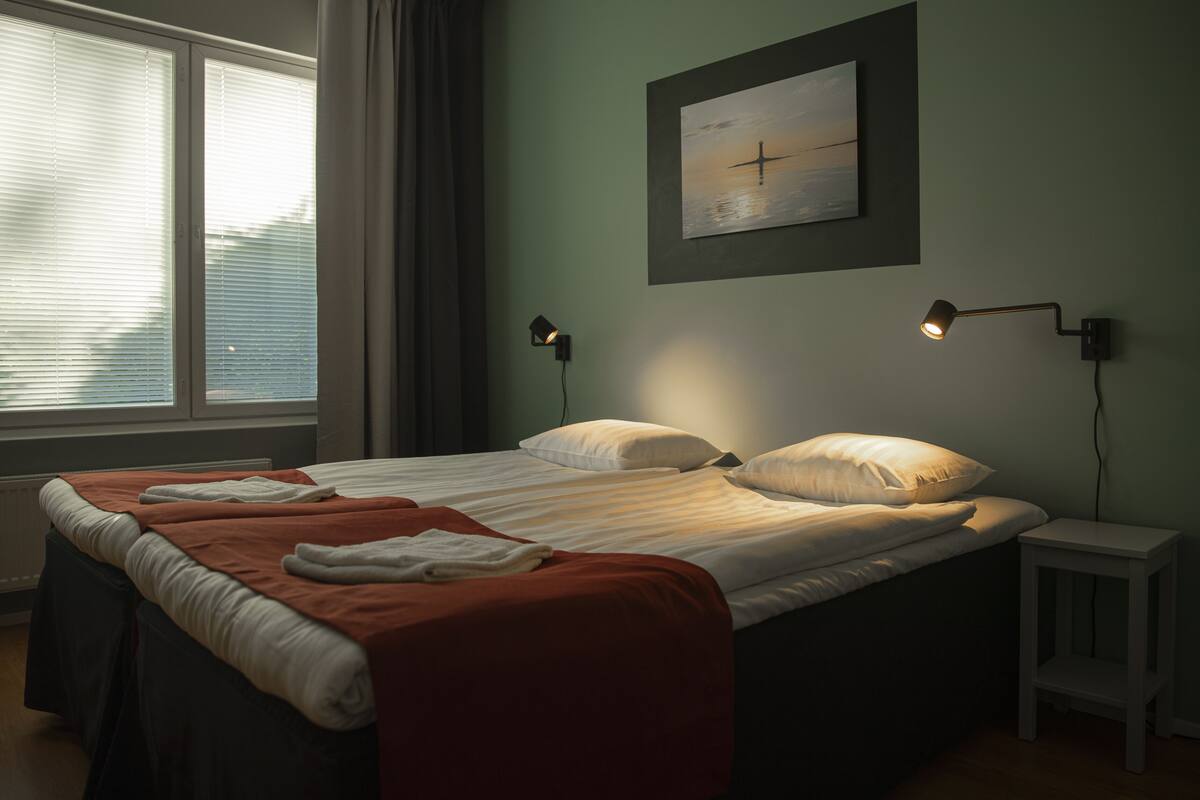 A neatly made bed with a red blanket and pillows, two bedside lamps, a small white nightstand, and a painting on the wall.
