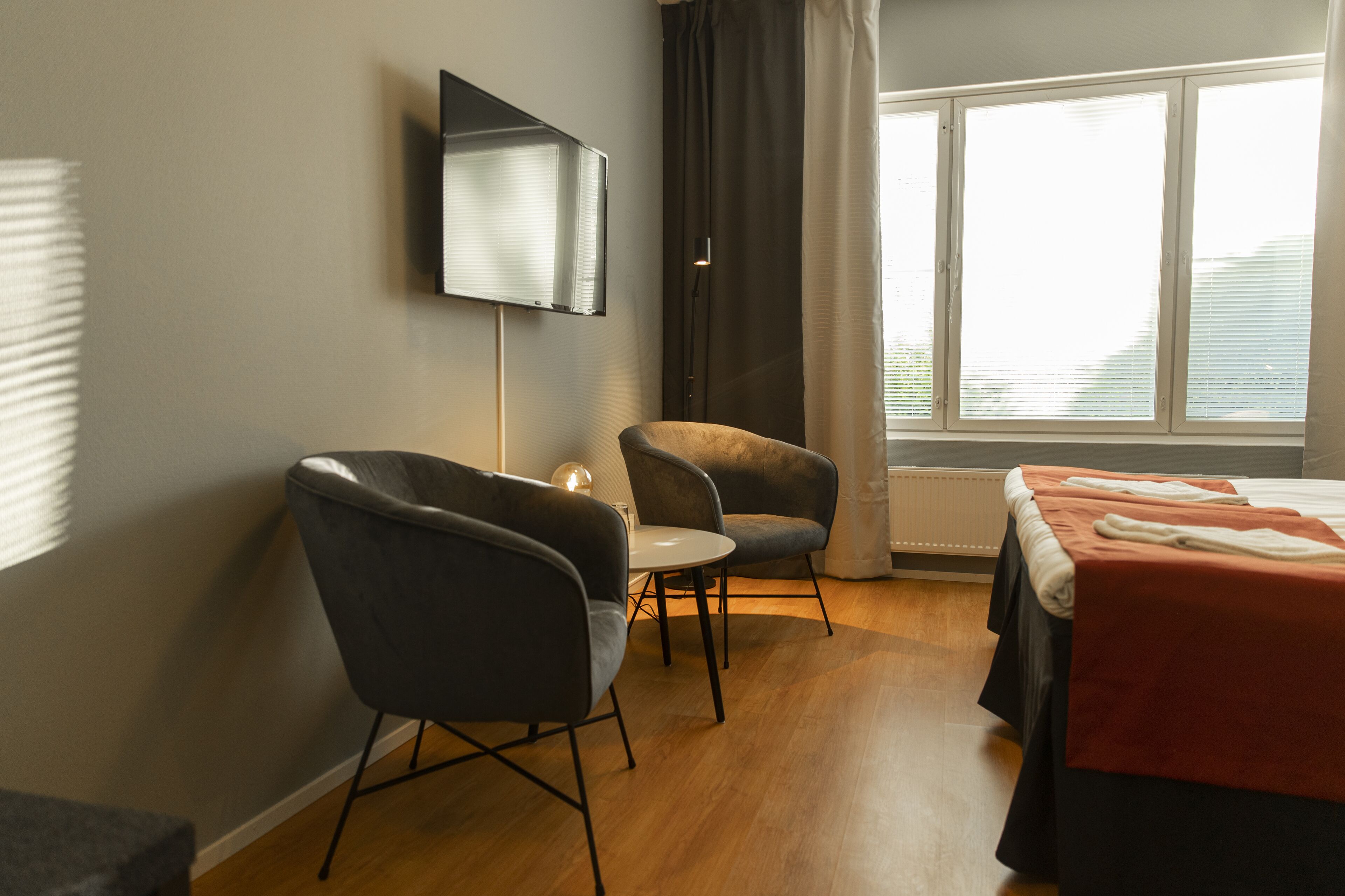 A hotel room with a TV mounted on the wall, two armchairs, a small table, a bed with a red blanket, and a large window with curtains.