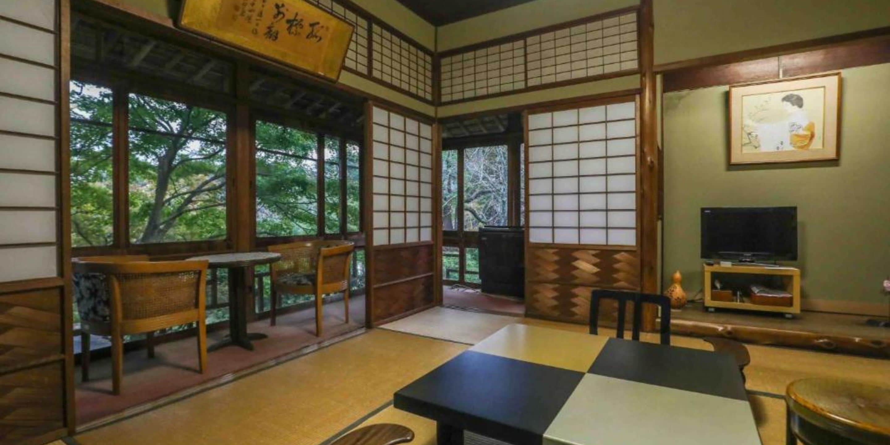 Een traditionele Japanse kamer met tatami vloeren, een lage tafel met stoelen, een televisie en een groot raam met uitzicht op bomen.