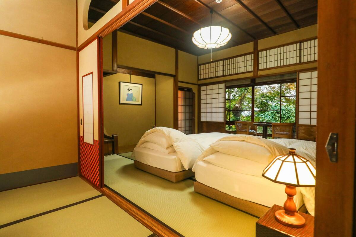 A traditional Japanese room with tatami flooring, a shoji screen, a bed with white bedding, and a nightstand with a lamp.