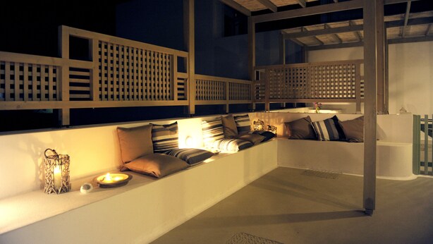 A well-lit outdoor seating area with a cushioned bench, a small table with a candle, and decorative items. - Almyris (Milos)