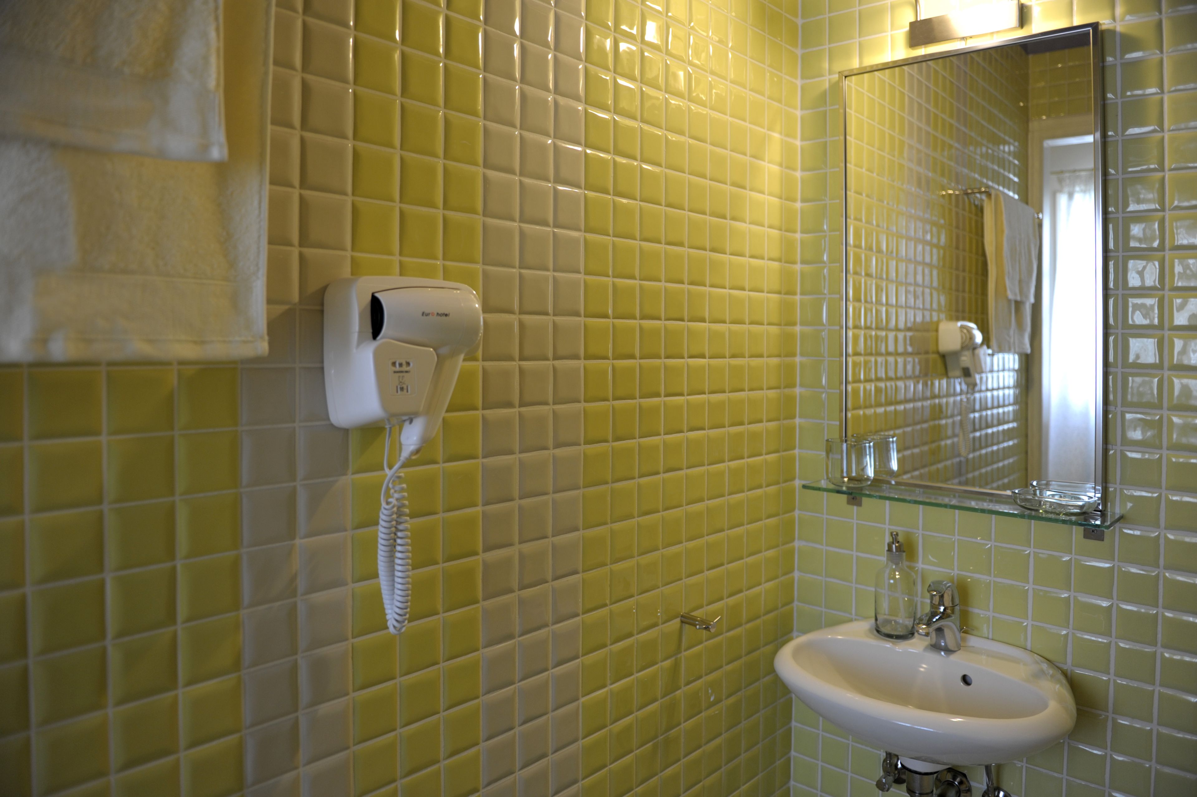 A bathroom with yellow tiled walls, a white sink, and a mirror with a glass shelf above it.