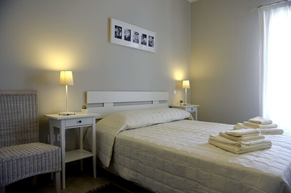 A neatly made bed with a white quilt, two bedside tables with lamps, a chair, and a framed photo on the wall.