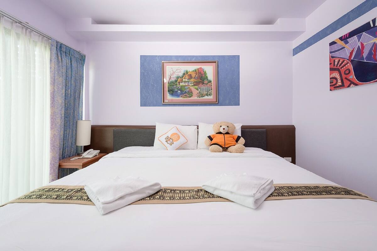 A neatly made bed with a teddy bear, a framed picture, and a lamp on a bedside table.