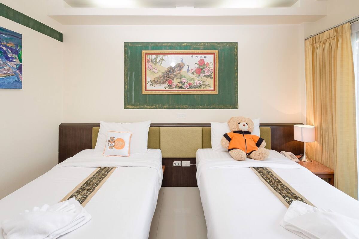 A hotel room with two beds, a teddy bear on one bed, a painting on the wall, and a lamp on the nightstand.