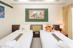 A hotel room with two beds, a teddy bear on one bed, a painting on the wall, and a lamp on the nightstand.