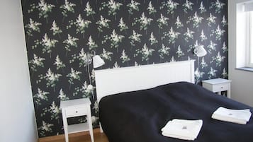 A bedroom with a dark floral wallpaper, a white bedside table, a white lamp, a bed with a dark cover, and folded towels on the bed.