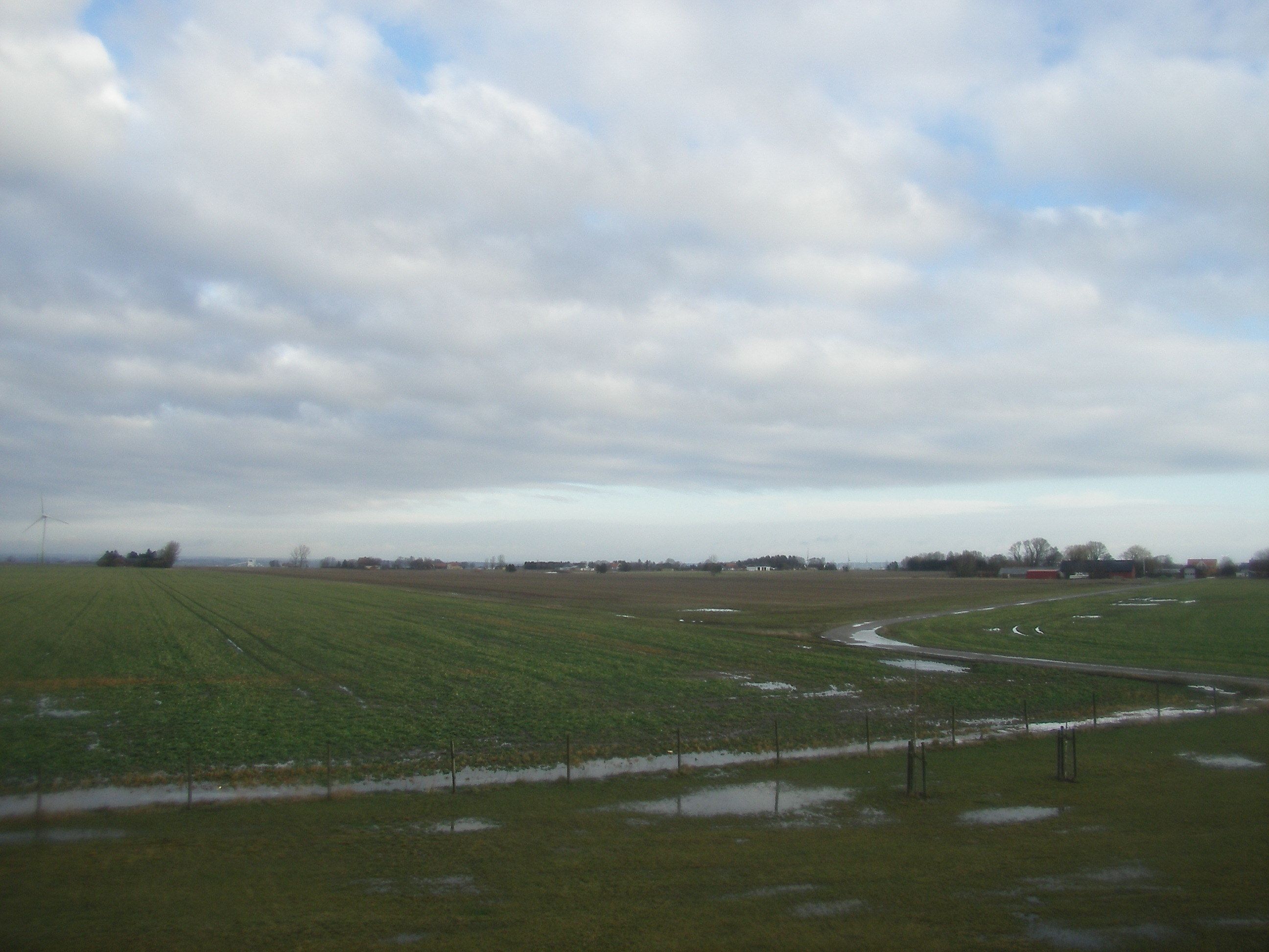 Paisaje rural con un campo, una pequeña masa de agua y edificios al fondo.