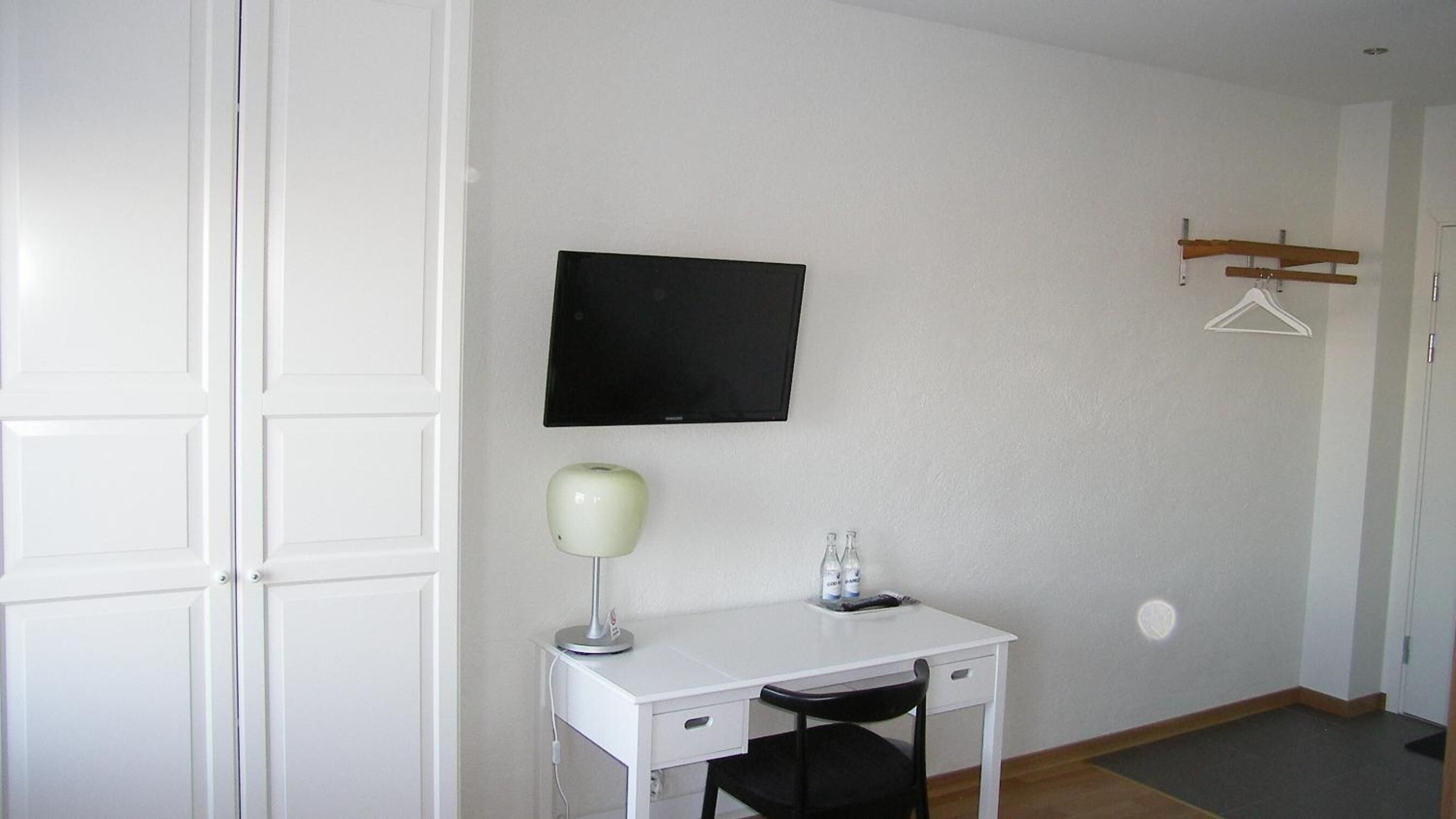 Una habitación pequeña y limpia con un escritorio blanco, una silla negra, un televisor montado en la pared, una lámpara de escritorio y un perchero.