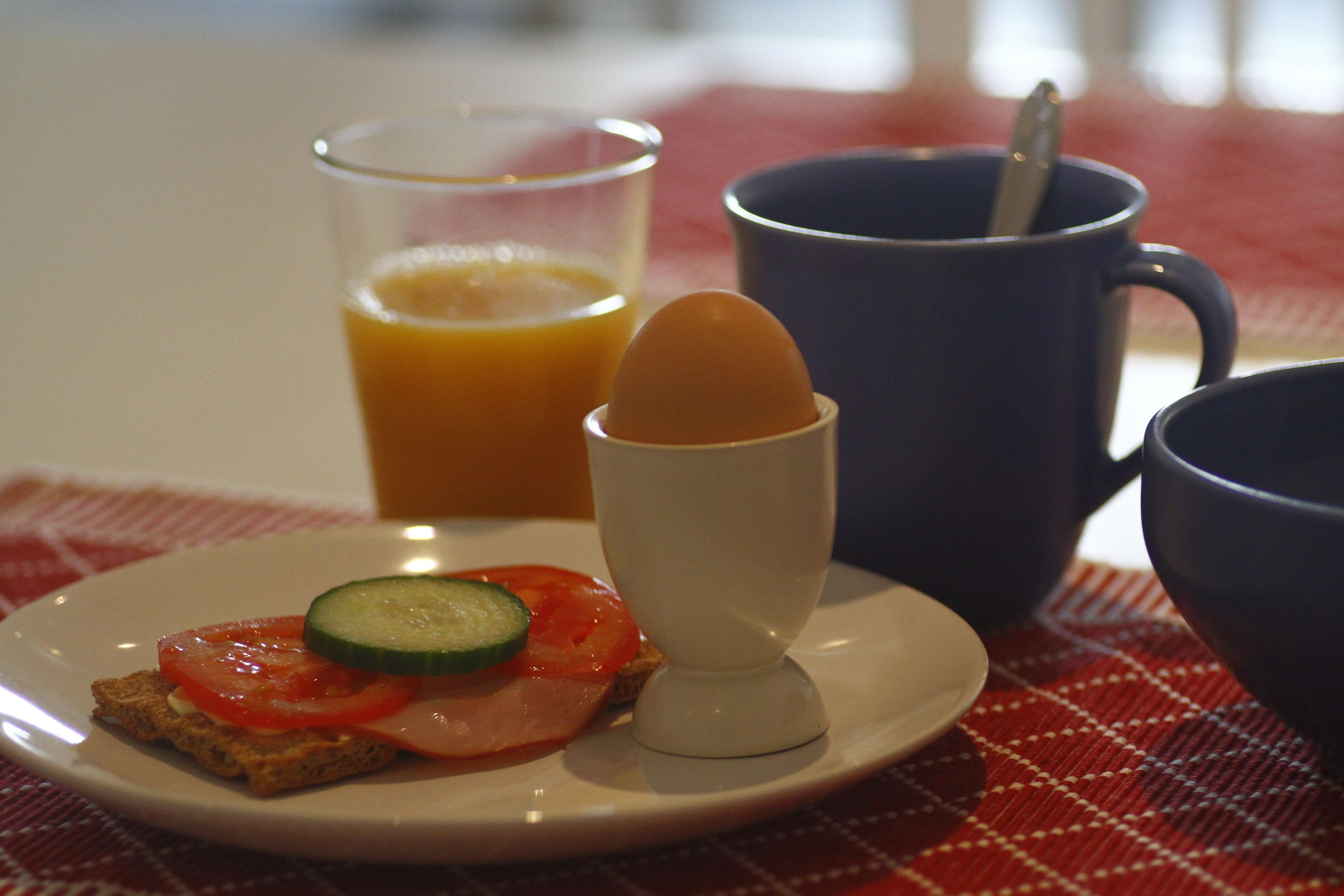 A breakfast plate with toast, tomato, cucumber, and a poached egg, accompanied by a glass of orange juice and a blue mug with a spoon.